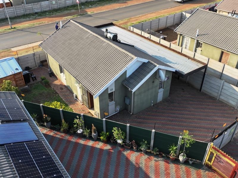 Aerial view of a residential property featuring a tiled roof, solar water heating system, paved driveway, carport, and secure perimeter walling.