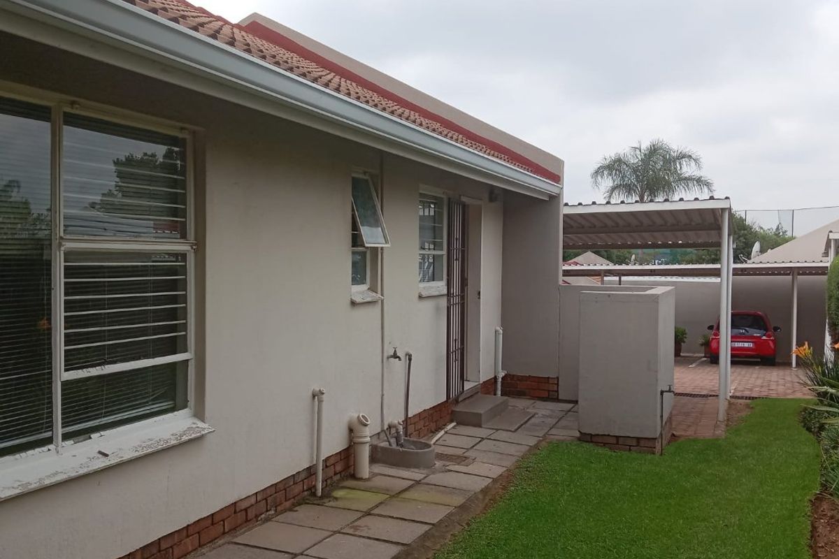 Rear exterior view of the apartment featuring a paved walkway, external water tap, and a covered carport area with a small lawn.