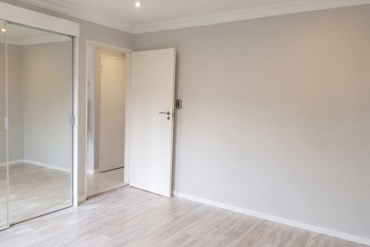 A bright, empty bedroom featuring light-toned laminate flooring, recessed downlighting, and a large built-in mirrored wardrobe.