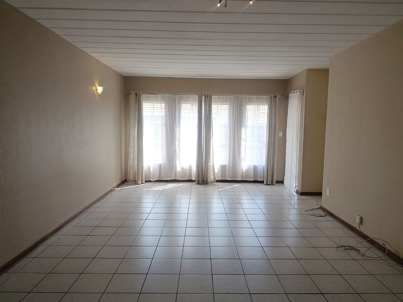 A spacious, empty living room featuring neutral tiled flooring, a panelled ceiling, and large windows allowing for ample natural light.