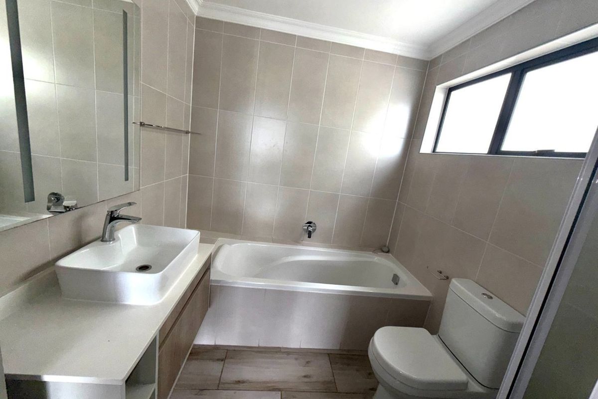 A clean, modern bathroom featuring a built-in vanity with a vessel sink, a bathtub, and a toilet, finished with neutral wall and floor tiles.