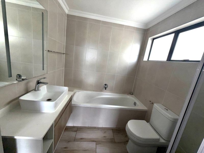 A clean, modern bathroom featuring a built-in vanity with a vessel sink, a bathtub, and a toilet, finished with neutral wall and floor tiles.