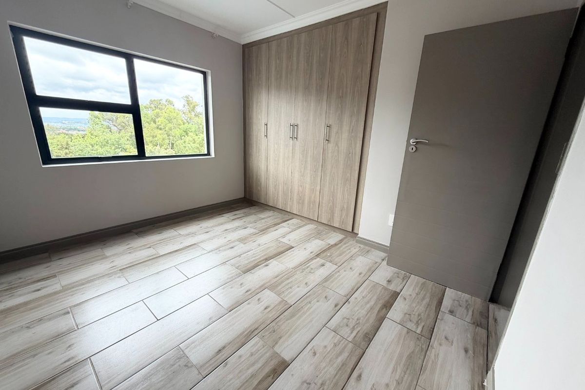 A bright, empty bedroom featuring modern wood-look tiled flooring, built-in cupboards, and a large window with views.