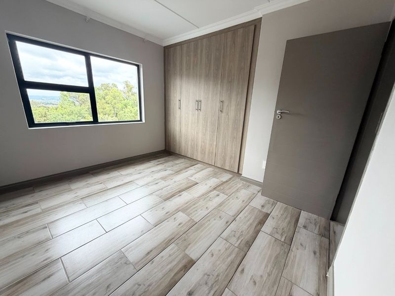 A bright, empty bedroom featuring modern wood-look tiled flooring, built-in cupboards, and a large window with views.