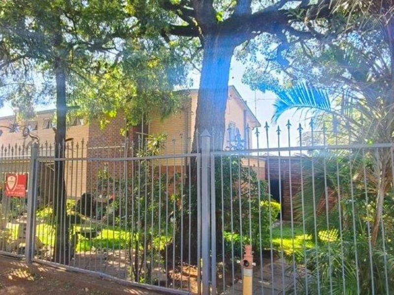 Front view of the property featuring a brick facade, mature trees, and secure metal perimeter fencing.