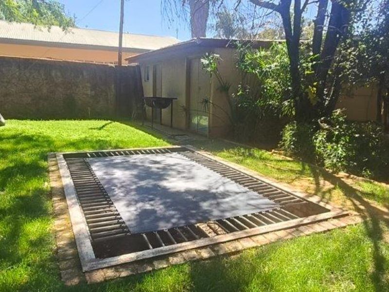 A private backyard garden featuring a sunken in-ground trampoline, established lawn, and a brick-walled boundary with an adjacent outbuilding.
