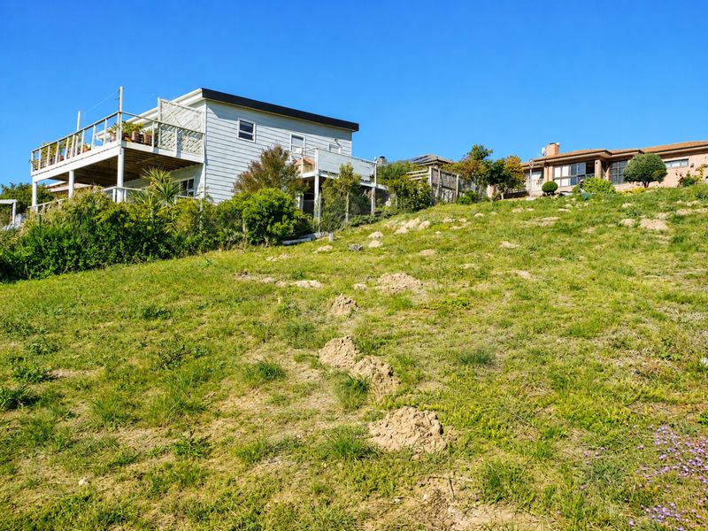 A view of a sloped residential vacant land plot with grass terrain, showing adjacent properties in the background.