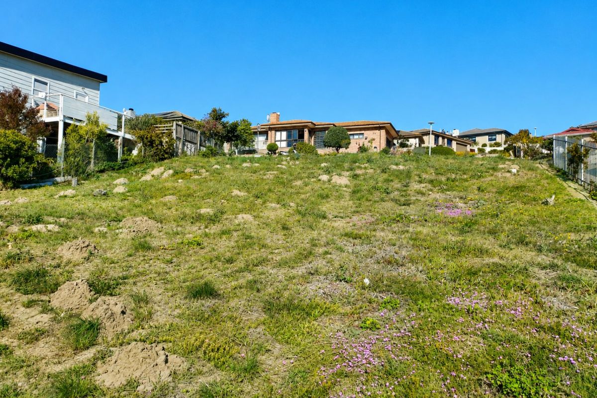 A view of a sloped, grass-covered vacant residential plot with surrounding boundary fencing and neighbouring properties visible in the background.