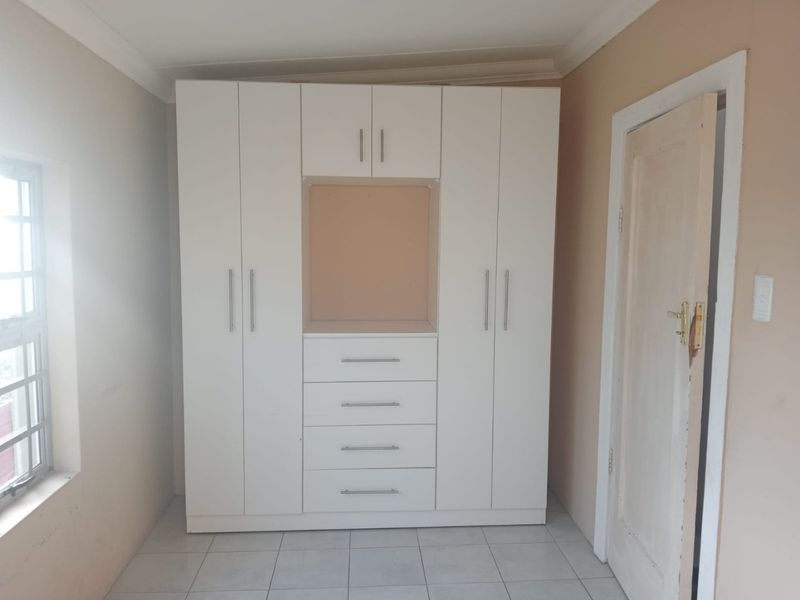 A bedroom featuring a large white built-in wardrobe with integrated drawers and shelving, tiled flooring, and a window with security bars.