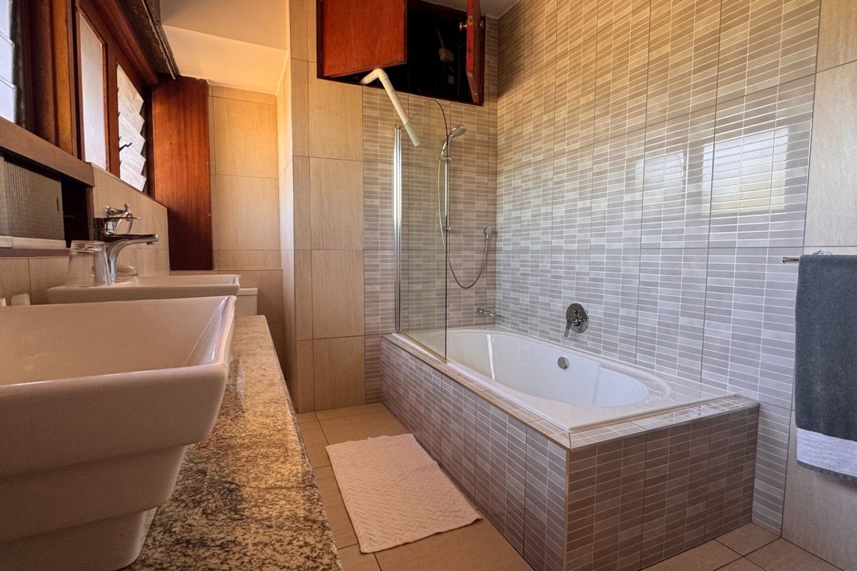 A functional bathroom featuring a double vanity with granite countertop, tiled walls, and a built-in bathtub with a glass shower screen.