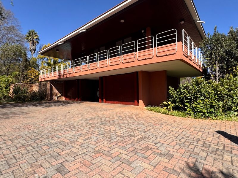 A modern architectural house featuring a prominent overhanging upper level with a balcony, set above a ground-level garage with a spacious paved driveway.