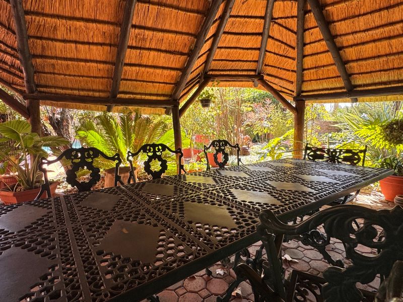 A covered outdoor patio area featuring a traditional thatched roof, timber support structure, and paved flooring, overlooking a lush garden setting.