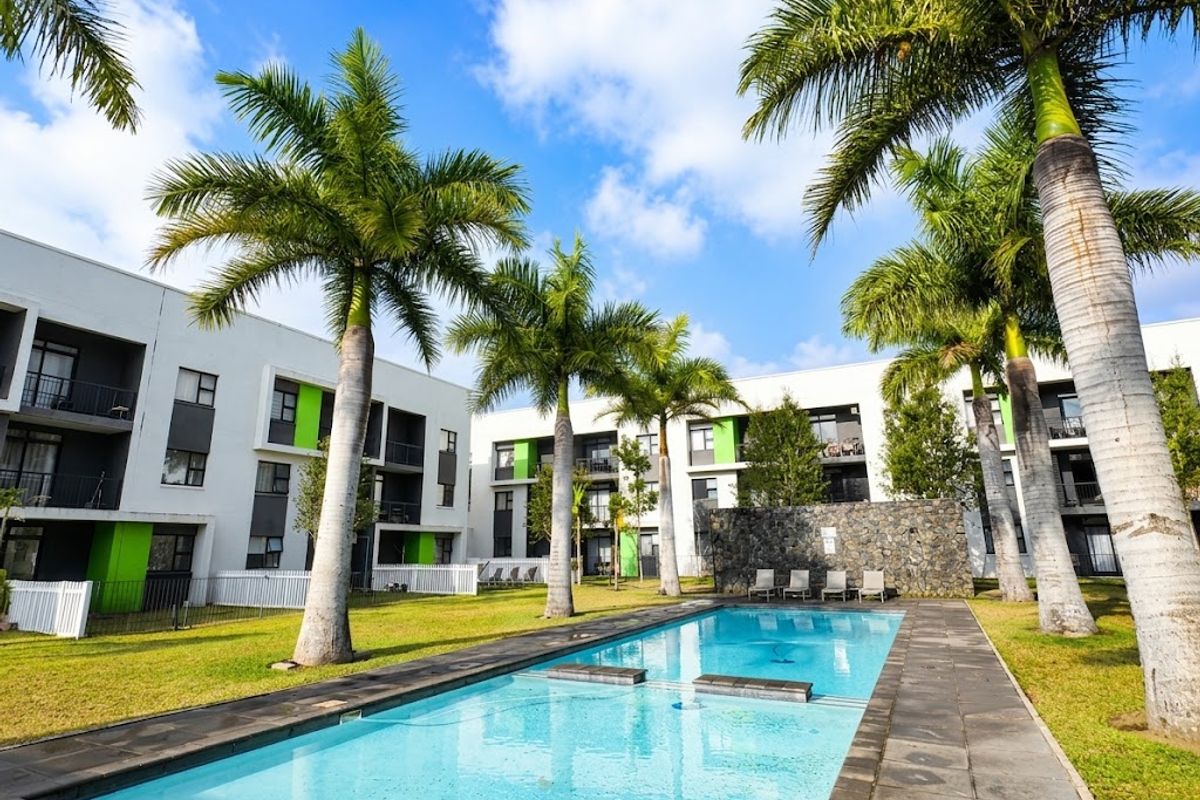 A well-maintained communal swimming pool area featuring a stone feature wall, paved decking, and landscaped gardens surrounded by modern apartment buildings.