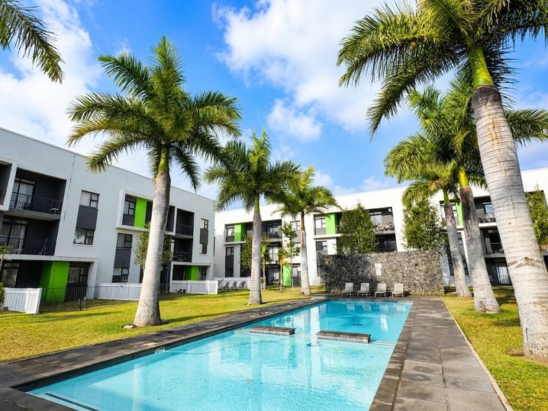 A well-maintained communal swimming pool area featuring a stone feature wall, paved decking, and landscaped gardens surrounded by modern apartment buildings.