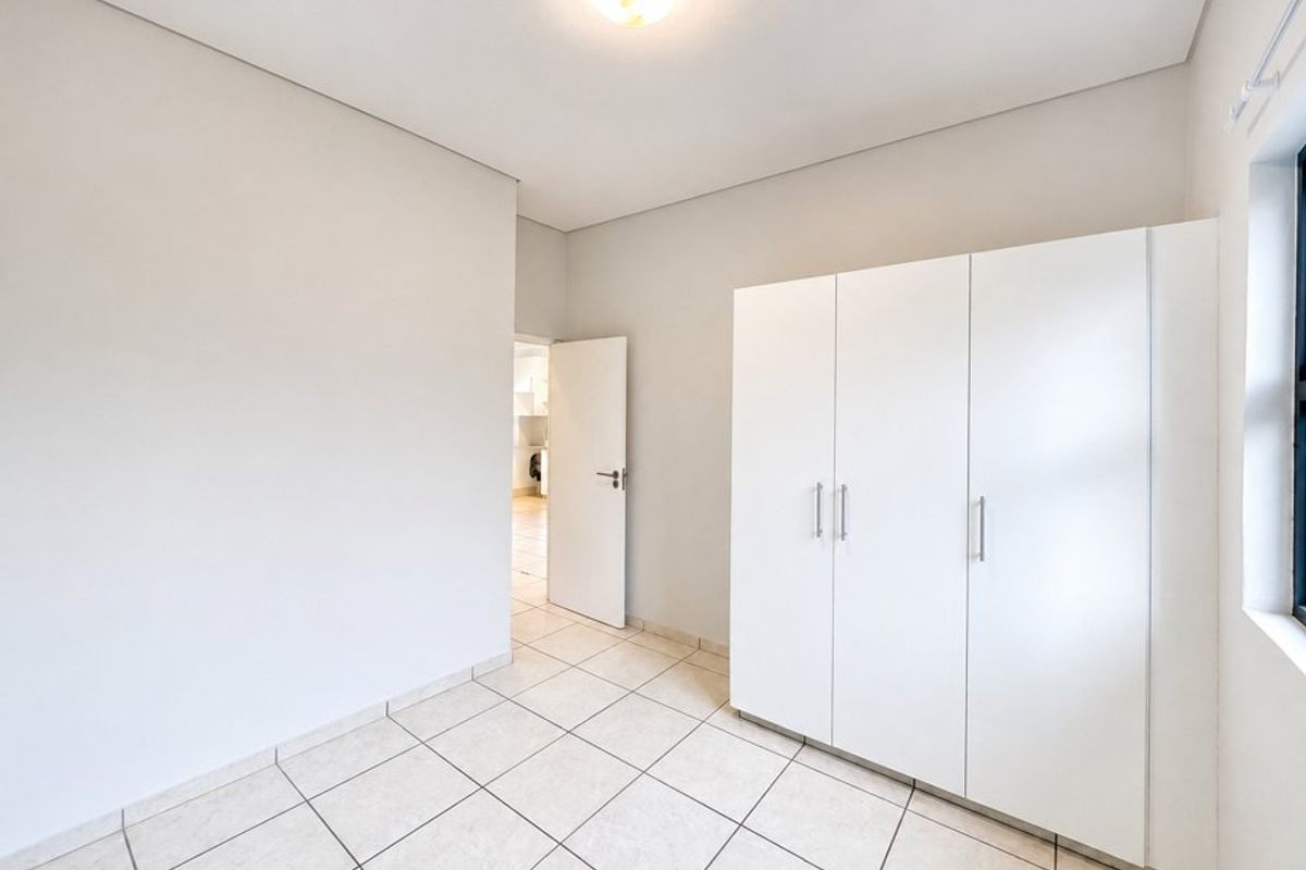 A bright, empty bedroom featuring neutral tiled flooring, a built-in white wardrobe, and a simple flush mount ceiling light fixture.