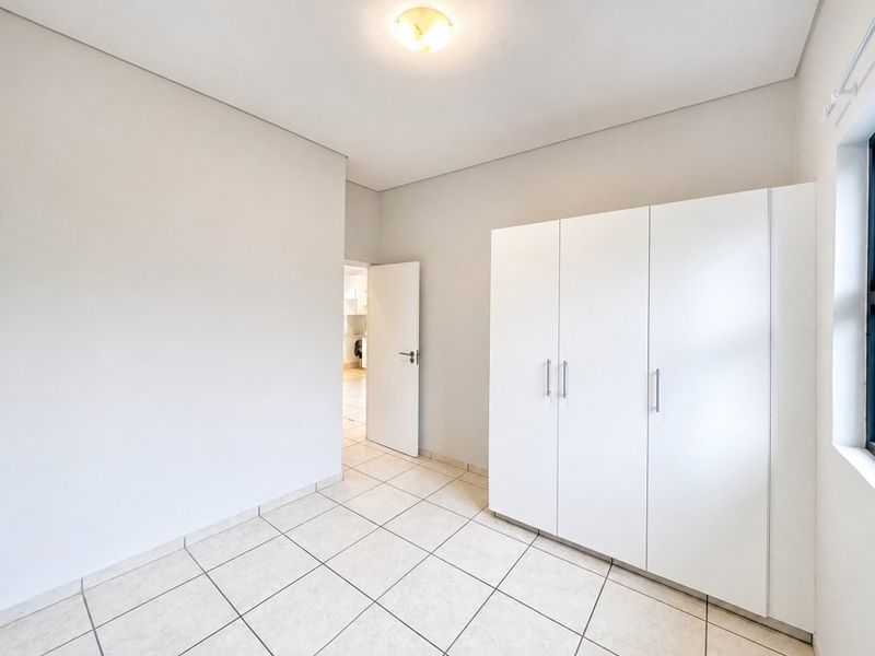 A bright, empty bedroom featuring neutral tiled flooring, a built-in white wardrobe, and a simple flush mount ceiling light fixture.