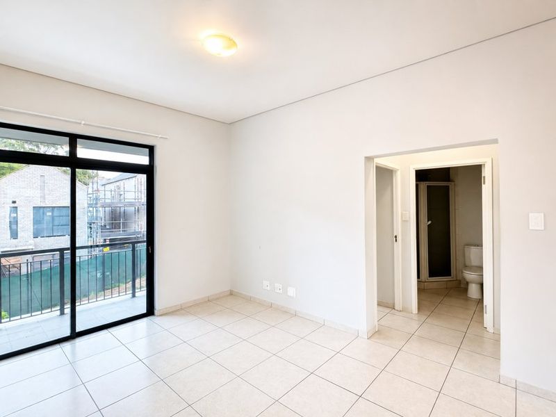 A bright, empty bedroom featuring tiled flooring, a sliding glass door leading to a balcony, and direct access to an en-suite bathroom.