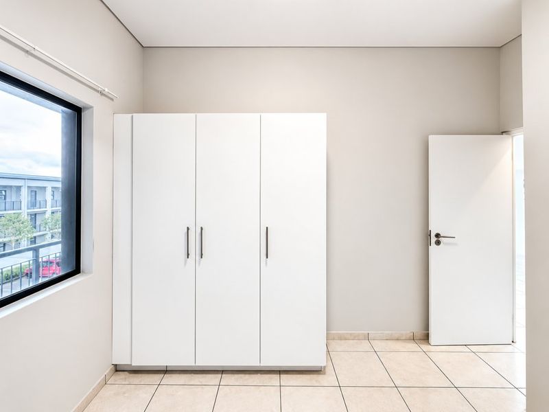 A bright, neutral bedroom featuring built-in white wardrobes, tiled flooring, and a large window providing natural light.