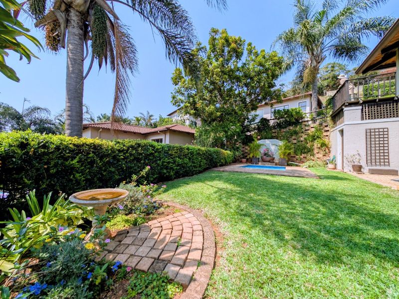 A well-maintained private garden featuring a small swimming pool, lush lawn, mature trees, and brick-paved pathways, set against a multi-level townhouse exterior.