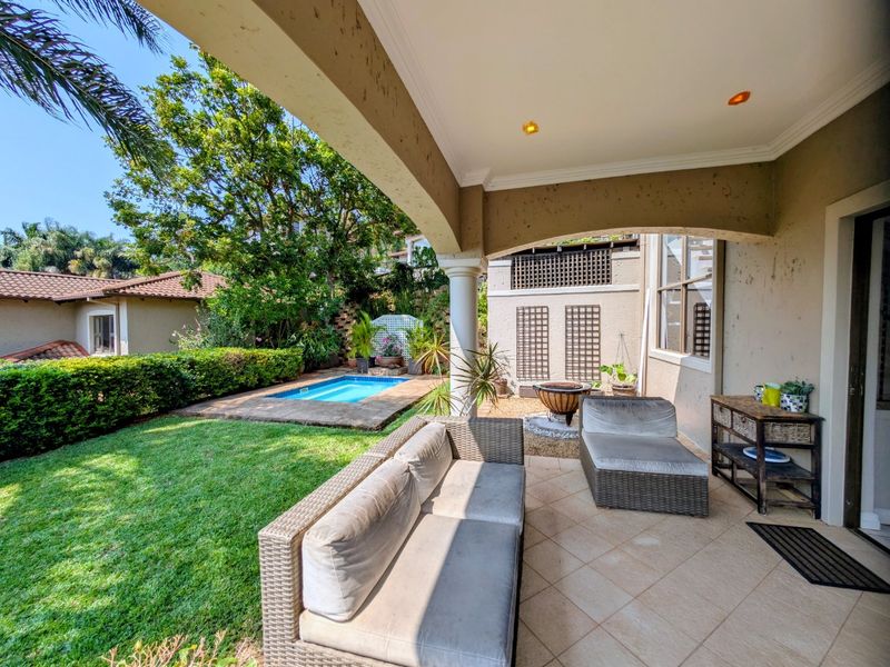 A covered patio area overlooking a private garden and swimming pool, featuring tiled flooring and recessed lighting.