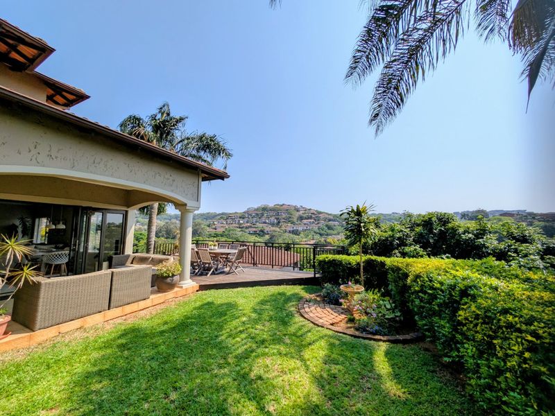 Rear exterior view featuring a covered patio, wooden deck, landscaped garden, and scenic hillside views.