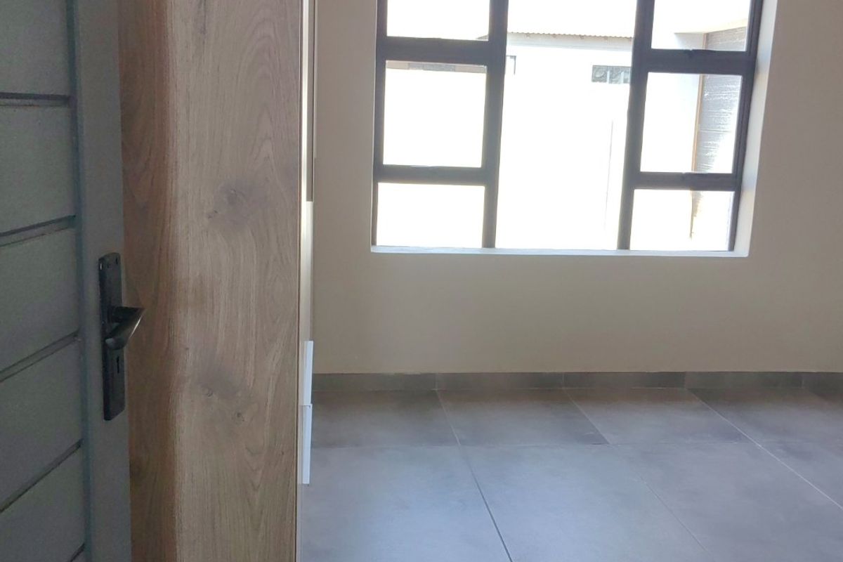 A bright, empty bedroom featuring modern grey floor tiles, built-in wooden cupboards, and large aluminium-framed windows.