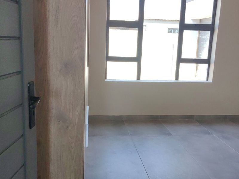 A bright, empty bedroom featuring modern grey floor tiles, built-in wooden cupboards, and large aluminium-framed windows.
