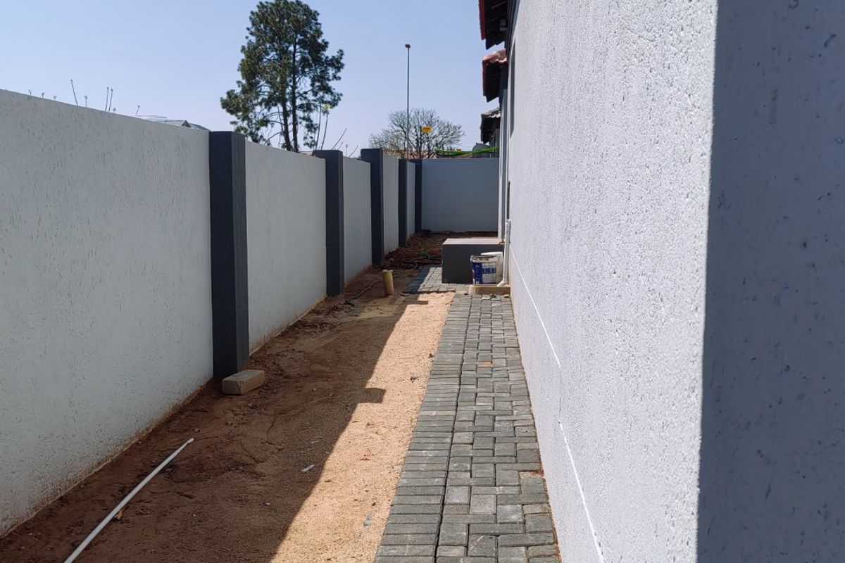 A side exterior view showing a paved walkway alongside the house, bordered by a high boundary wall and unfinished garden area.