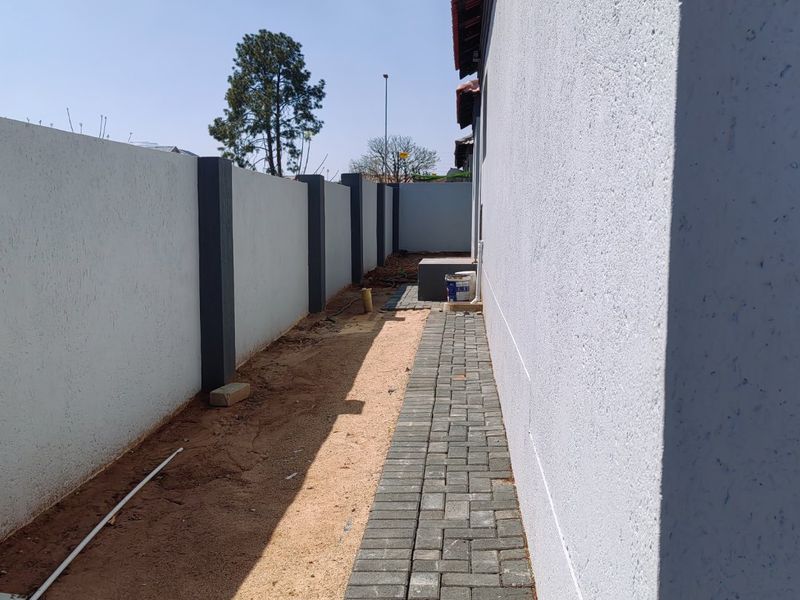 A side exterior view showing a paved walkway alongside the house, bordered by a high boundary wall and unfinished garden area.