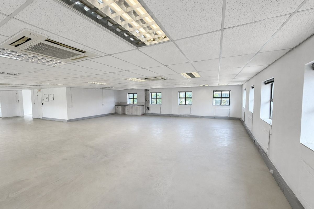 A spacious, open-plan commercial office area featuring a suspended ceiling, recessed lighting, integrated air conditioning, and a small kitchenette corner.
