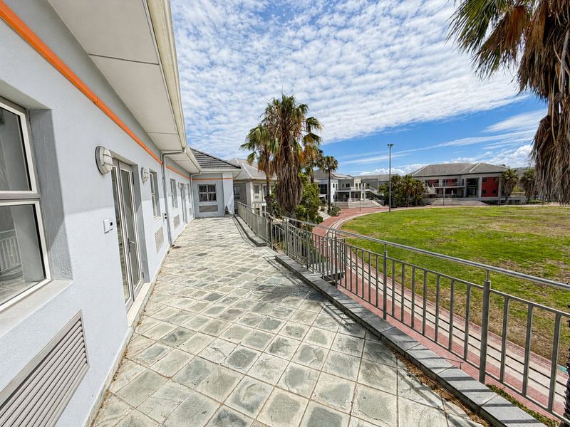 A spacious elevated balcony featuring stone tiling and metal railings, overlooking a landscaped commercial courtyard and green space.