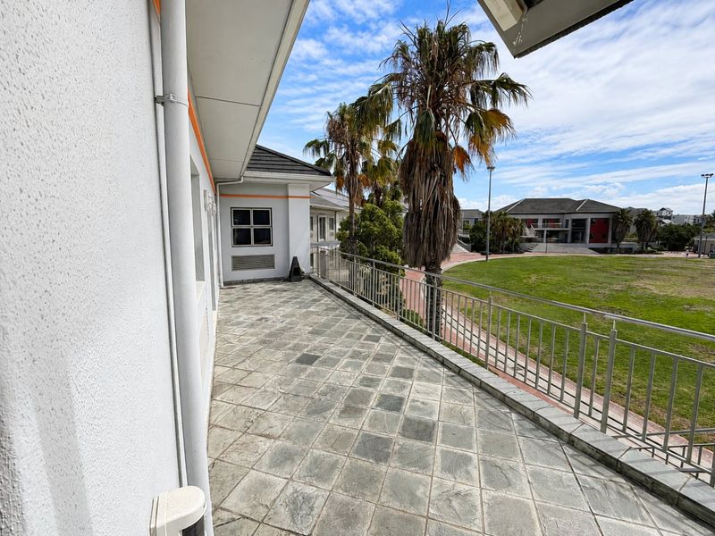 A spacious covered balcony featuring stone tiled flooring and a metal balustrade, overlooking a landscaped commercial estate area.
