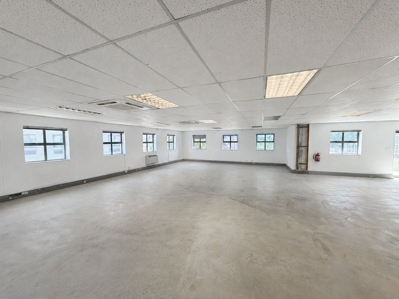 Spacious open-plan commercial office space featuring concrete flooring, suspended ceilings with recessed lighting, and multiple windows providing natural light.