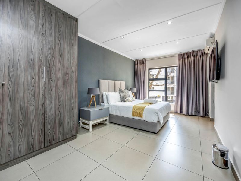 A well-maintained bedroom featuring large built-in cupboards, neutral tiled flooring, recessed lighting, and an air conditioning unit for climate control.