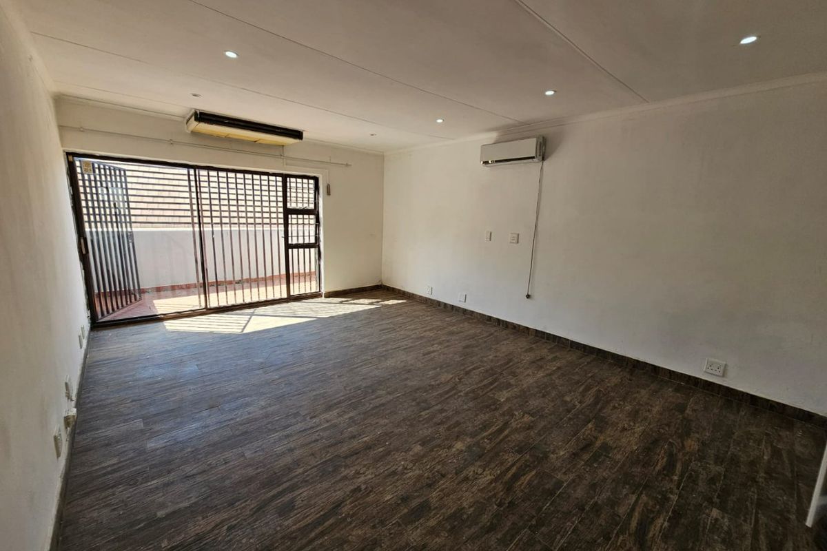 An empty, open-plan living area featuring dark wood-look floor tiles, recessed lighting, a wall-mounted air conditioning unit, and access to an outdoor space.