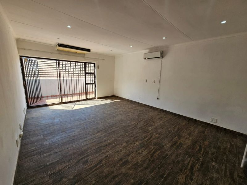 An empty, open-plan living area featuring dark wood-look floor tiles, recessed lighting, a wall-mounted air conditioning unit, and access to an outdoor space.