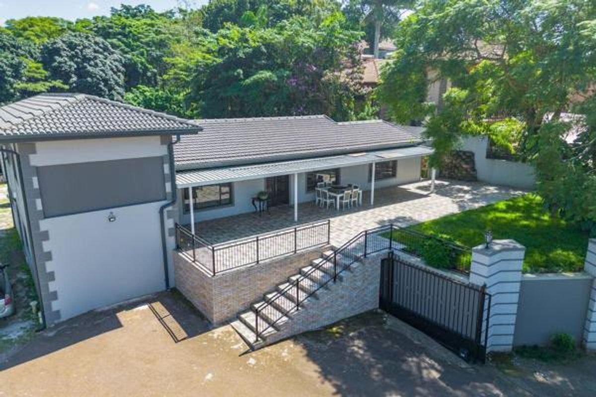 Aerial view of the rear exterior featuring a spacious covered patio, brick staircase, paved driveway, and secure boundary wall with gate.