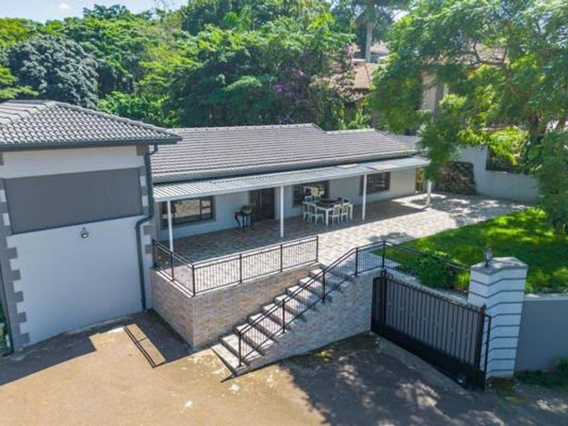 Aerial view of the rear exterior featuring a spacious covered patio, brick staircase, paved driveway, and secure boundary wall with gate.