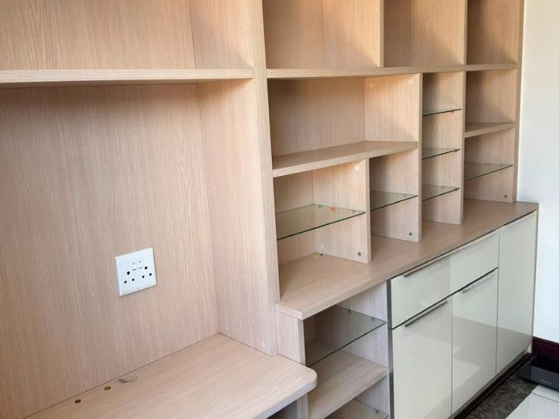 A modern, custom-built light wood office unit featuring extensive shelving, integrated desk space, and sleek lower cabinetry with glass accents.