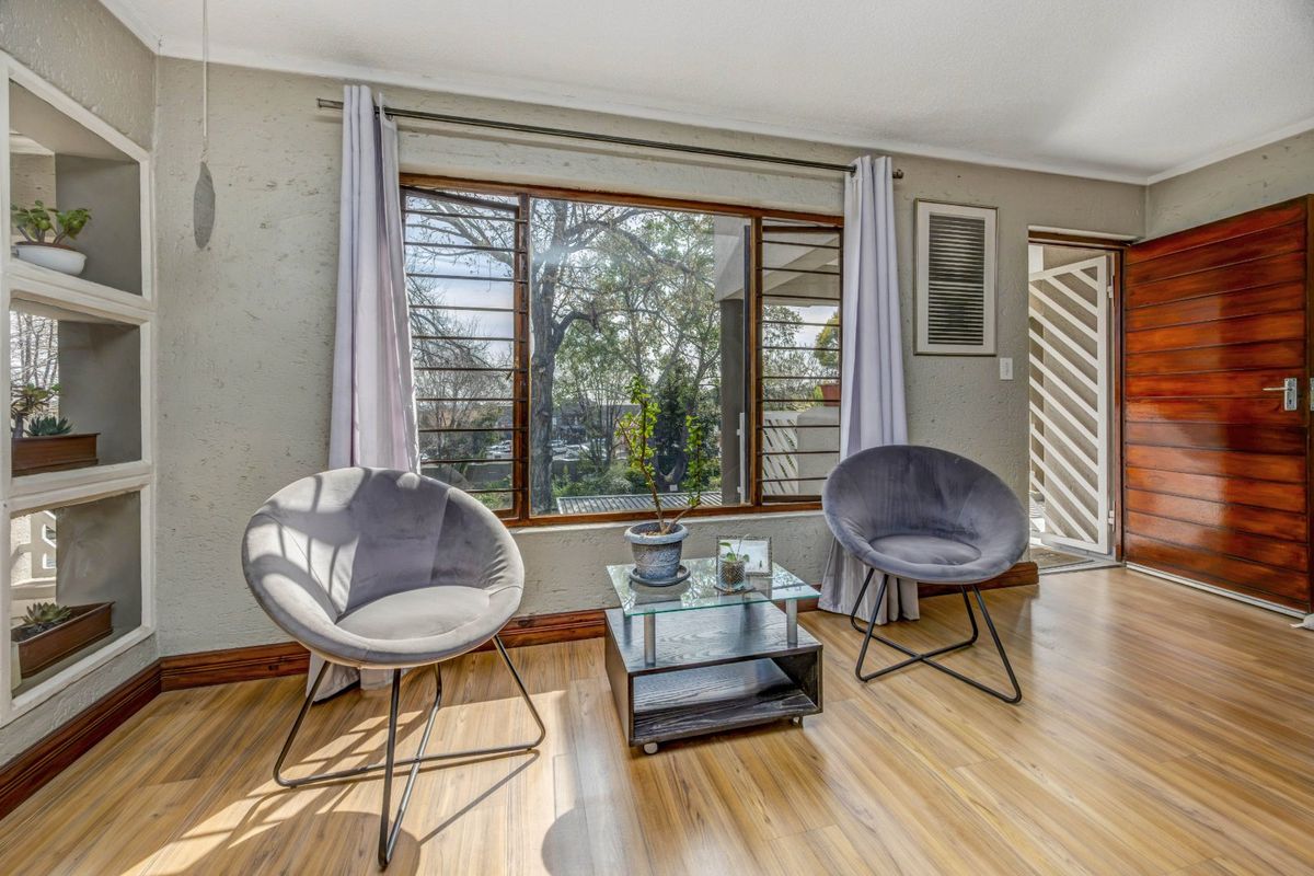 A bright living area featuring laminate flooring, built-in wall shelving, and a large window with security bars, leading to a timber entrance door.