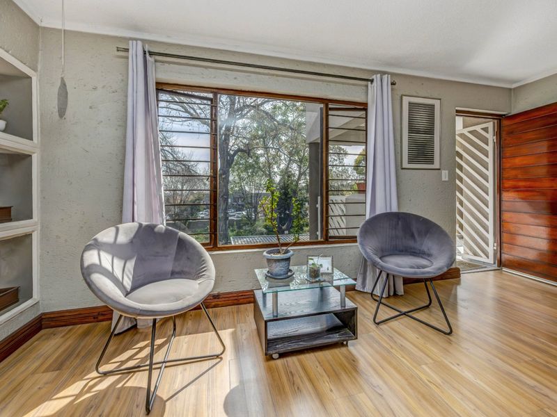 A bright living area featuring laminate flooring, built-in wall shelving, and a large window with security bars, leading to a timber entrance door.