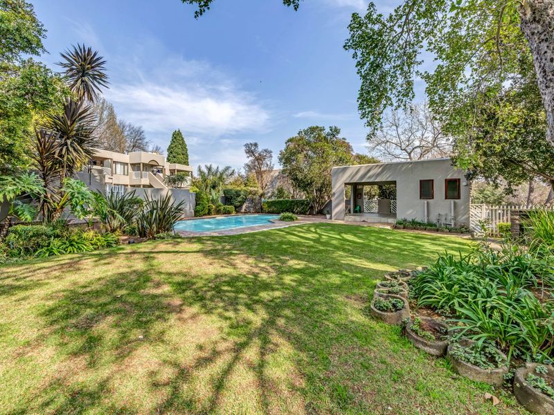 A well-maintained communal garden area featuring a swimming pool and a covered outdoor structure, surrounded by mature trees and established landscaping.