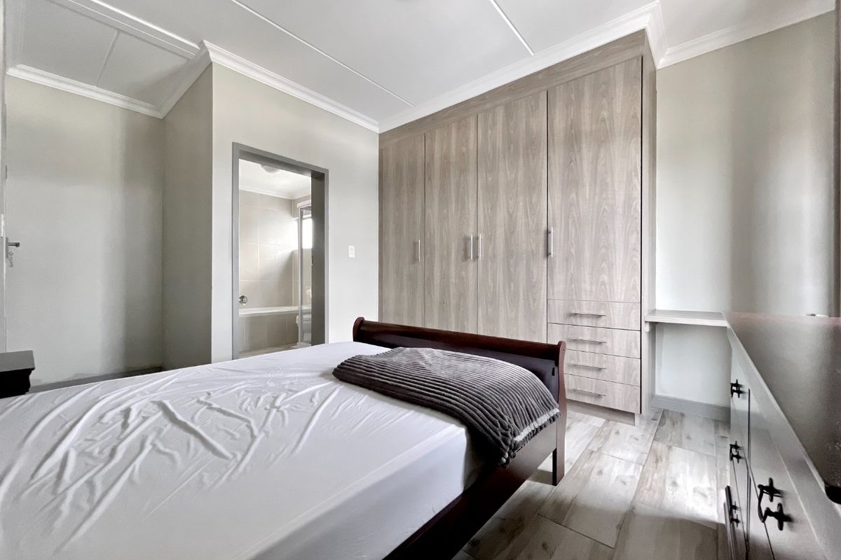 A well-maintained bedroom featuring modern wood-grain built-in cupboards, integrated vanity desk, and neutral tiled flooring with direct access to an en-suite bathroom.