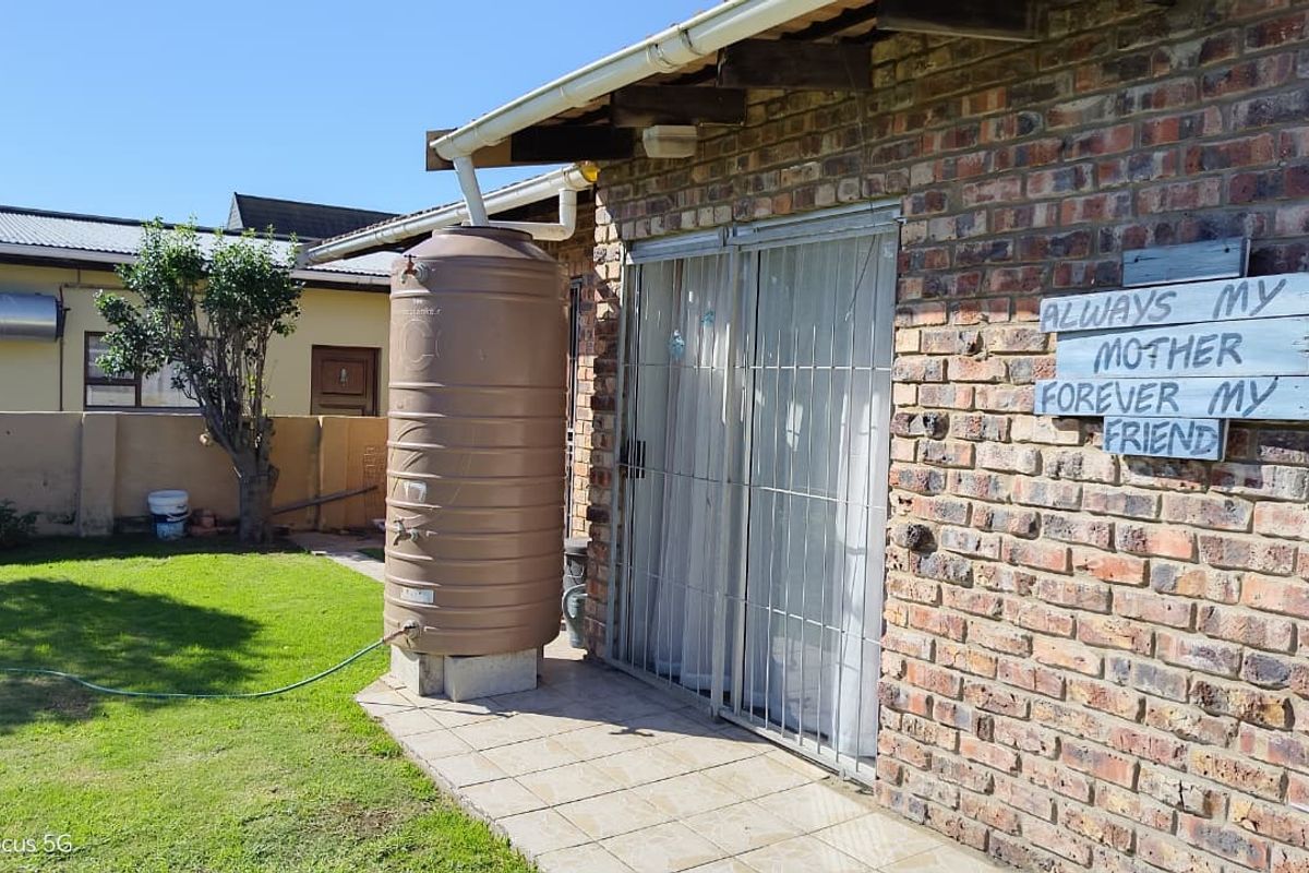 Rear exterior view featuring a face brick wall, sliding glass door with security gate, tiled patio area, and a water storage tank on the lawn.