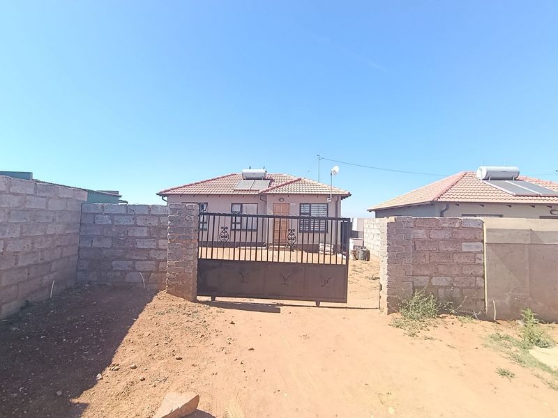 Front view of a single-story house featuring a tiled roof, solar water heater, and a secure metal gate with an unpaved yard.