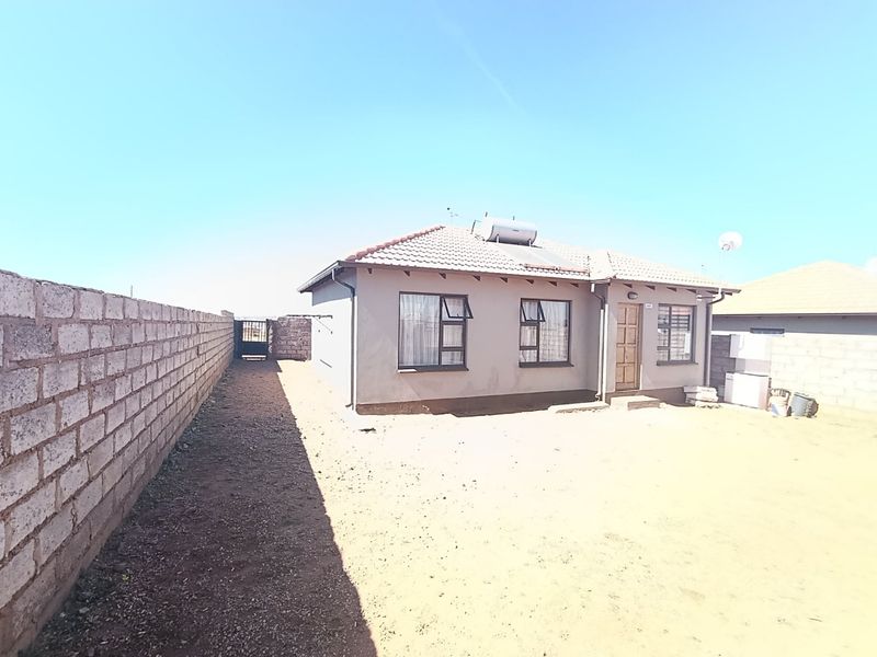 A single-story residential house featuring a tiled roof, solar water heater, and a large, unpaved yard enclosed by a boundary wall.