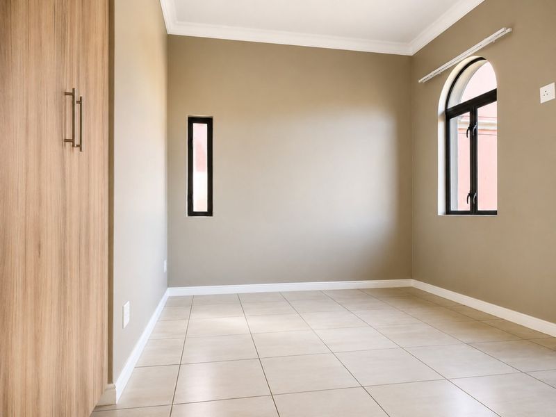 A bright, empty bedroom featuring neutral tiled flooring, built-in wooden cabinetry, and unique arched and vertical window detailing.
