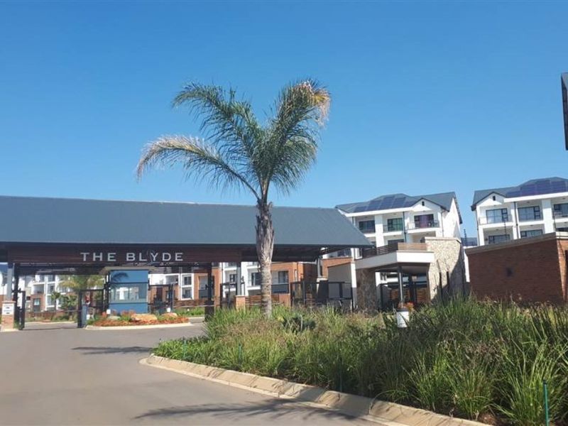 The entrance to The Blyde apartment complex, featuring a modern security gatehouse, paved driveway, and contemporary multi-story residential buildings with brick and white finishes.