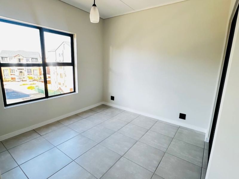 A bright, empty bedroom featuring neutral tiled flooring, a modern pendant light, and large aluminium-framed windows providing natural light.