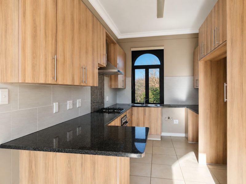 Modern kitchen featuring light wood cabinetry, black granite countertops, built-in gas hob, oven, extractor fan, and tiled finishes with an arched window.